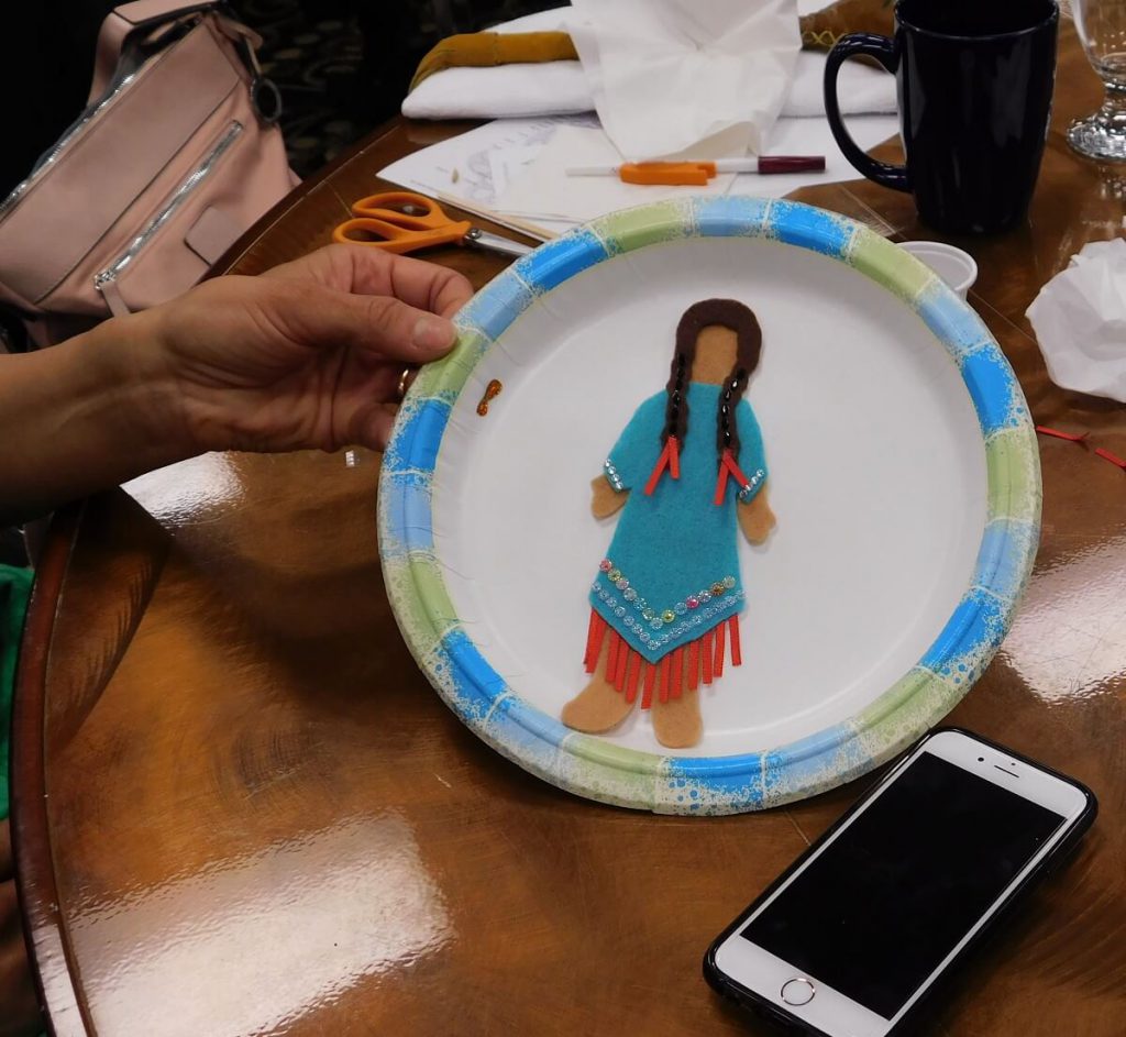 felt doll on plate with blue dress, red ribbons hanging down, and ribbons in hair