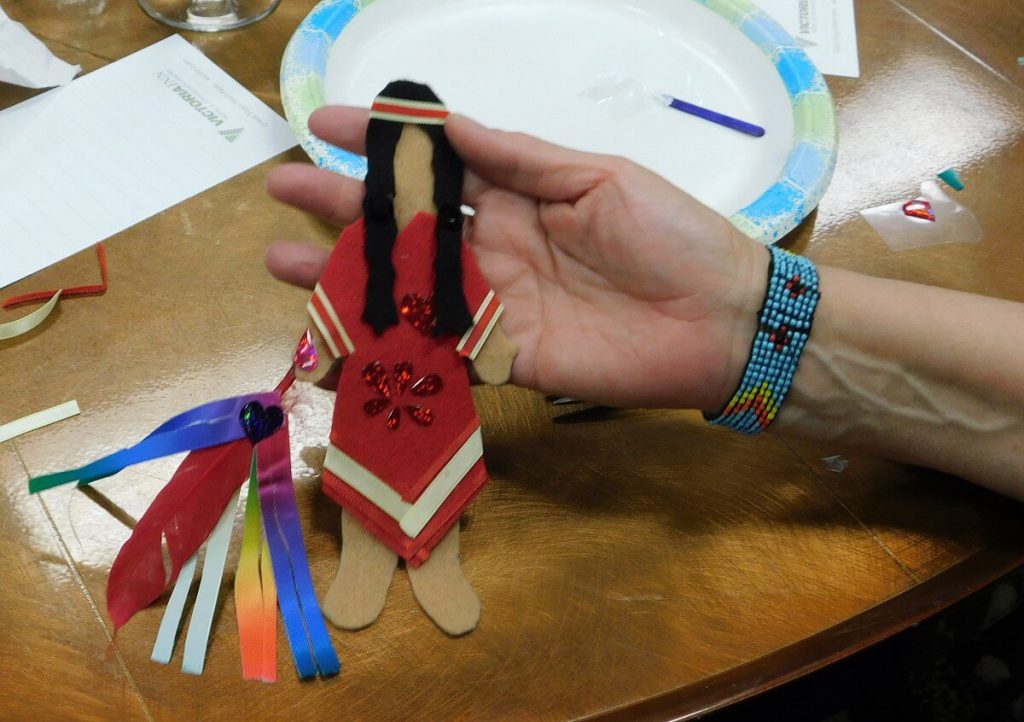 felt doll wearing red ribbon dress, holding a feather with ribbons attached