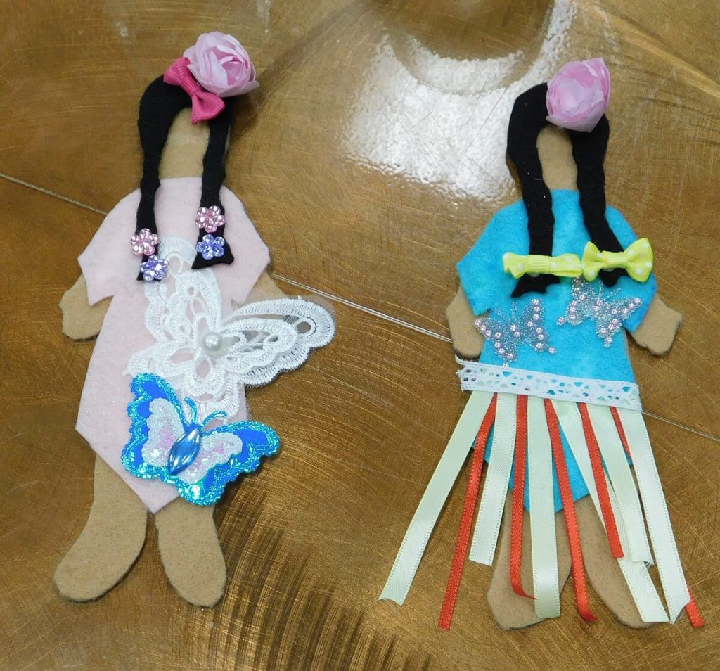 felt doll on left with flower on head and pink dress with butterflies, and a felt doll wearing a blue ribbon dress