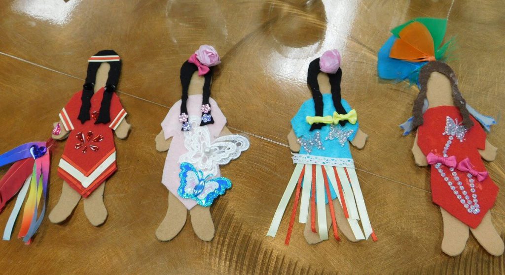 felt dolls from left to right" doll in red dress holding a ribboned feather, doll in light pink and butterflies on dress, doll in a blue ribbon dress, and a doll in a red dress with feathers on head