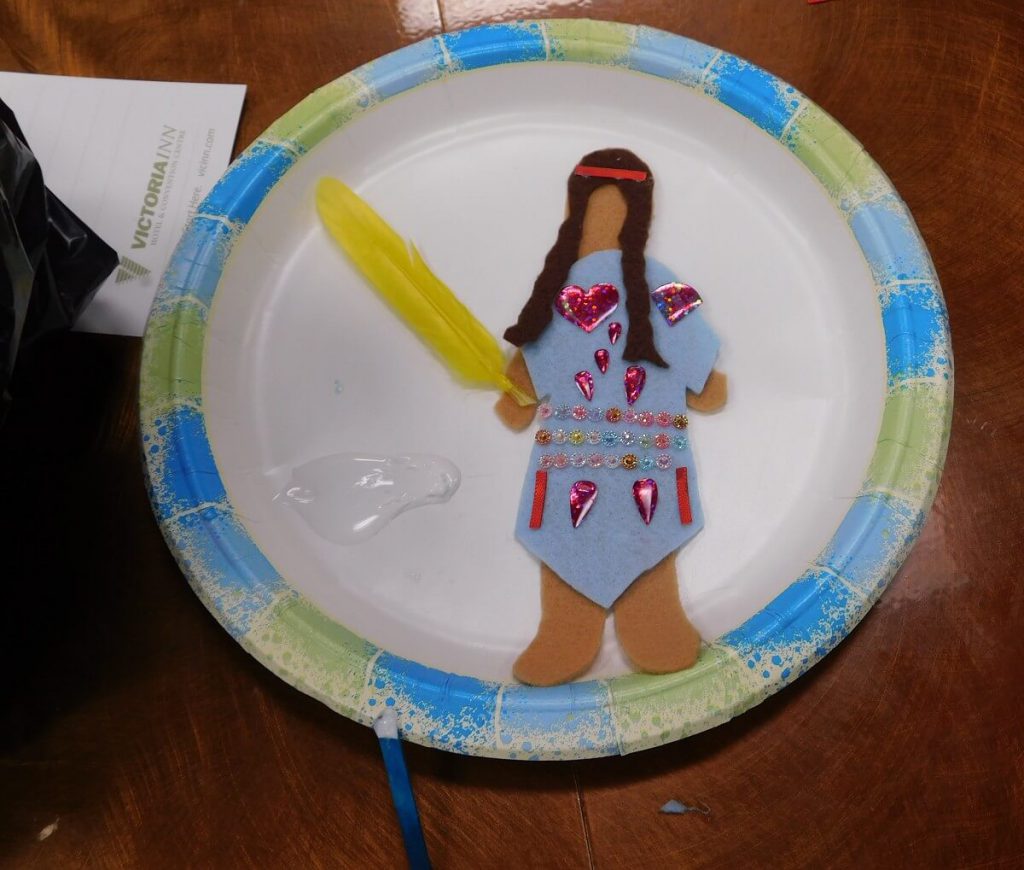 felt doll on a plate wearing a blue dress with a yellow feather in hand