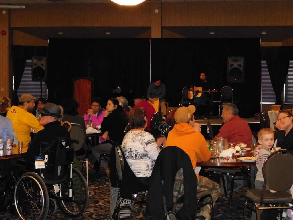 group of people in large room sitting at tables with performers playing guitar and drumming in background