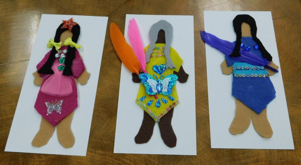 three felt dolls on paper strips, from left to right: felt doll in pink with yellow bows in hair, dolll in yellow with butterflies holding two feathers, and doll in blue holding feather